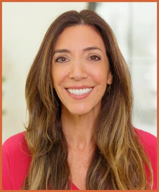 Image of Nicole Johnson, Co-founder of the FOXG1 Research Foundation,  with long, wavy brown hair and a bright smile, wearing a red top, in an indoor setting.