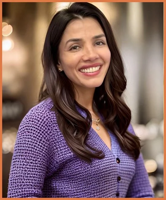 A smiling woman with long dark hair wearing a purple textured sweater and jewelry, standing indoors with blurred background lighting.