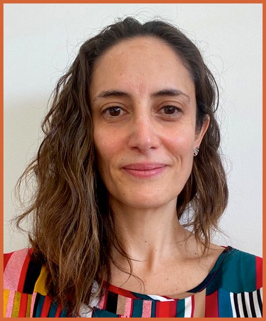 A woman with wavy brown hair and earrings, smiling slightly, wearing a colorful, striped top, standing against a plain light background.