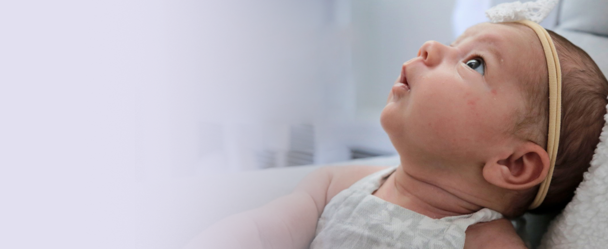 A baby with FOXG1 syndrome lying on a pillow, looking upwards with curiosity, wearing a beige headband and a white sleeveless top.