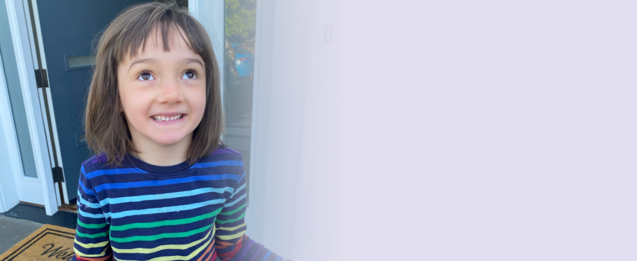 A young girl with shoulder-length brown hair, wearing a rainbow-striped long-sleeve shirt, smiling and looking up, standing outside near a door.