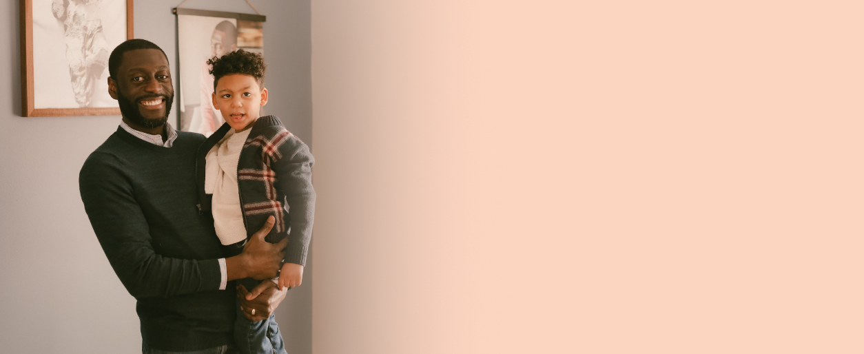 A man with a beard and a young boy with FOXG1 syndrome smiling, standing indoors against a light-colored wall with artwork.