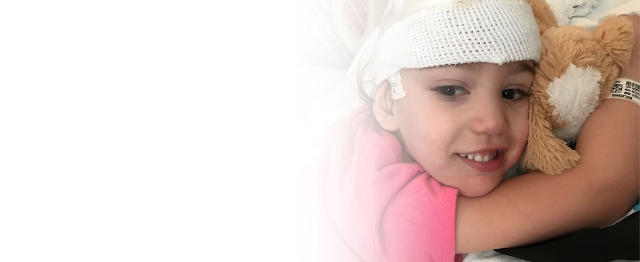 Young girl named Josie with FOXG1 syndrome  in hospital bed with a bandage on her head, smiling and hugging a teddy bear.