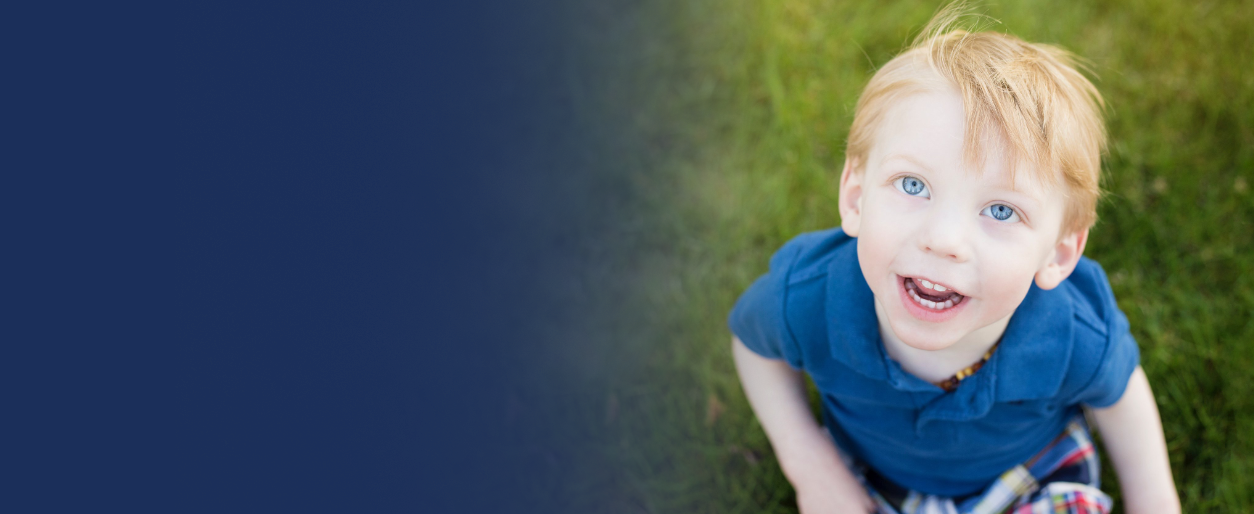 A young boy with blond hair and blue eyes smiling and looking up at the camera, sitting on green grass.