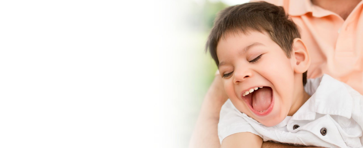 A young boy laughing happily with his eyes closed, sitting on someone's lap outdoors.