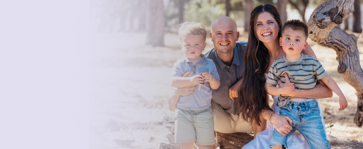 A family of four, including a woman, a man, and two young boys, smiling and sitting outdoors under a tree on a sunny day.