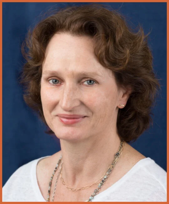 Headshot of a woman with short, wavy brown hair, blue eyes, wearing a white top and layered necklaces, against a blue background.