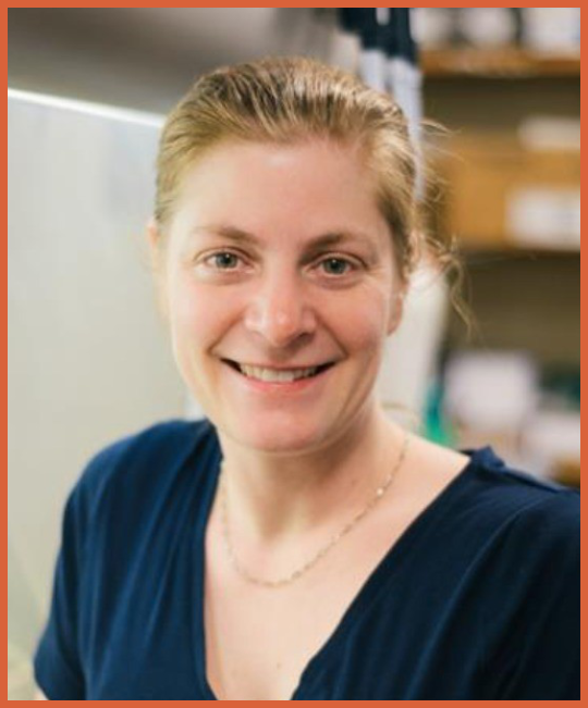 Smiling woman with blonde hair in a blue shirt, standing indoors with shelves in the background.