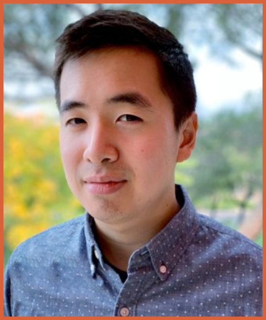 A young Asian man with short black hair and a slight smile, wearing a blue, button-up shirt with a subtle pattern, standing outdoors with blurred trees in the background.