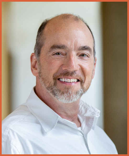 Portrait of a middle-aged man with short hair, a beard, and a broad smile, wearing a white collared shirt.