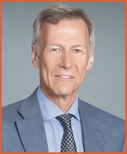 A professional headshot of an older man with gray hair wearing a blue suit, light blue dress shirt, and patterned tie, against a plain gray background.