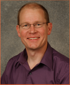 A smiling man with glasses wearing a purple shirt, posed against a gray background.