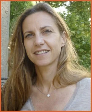 Woman with long light brown hair outdoors, smiling, with trees and greenery in background.