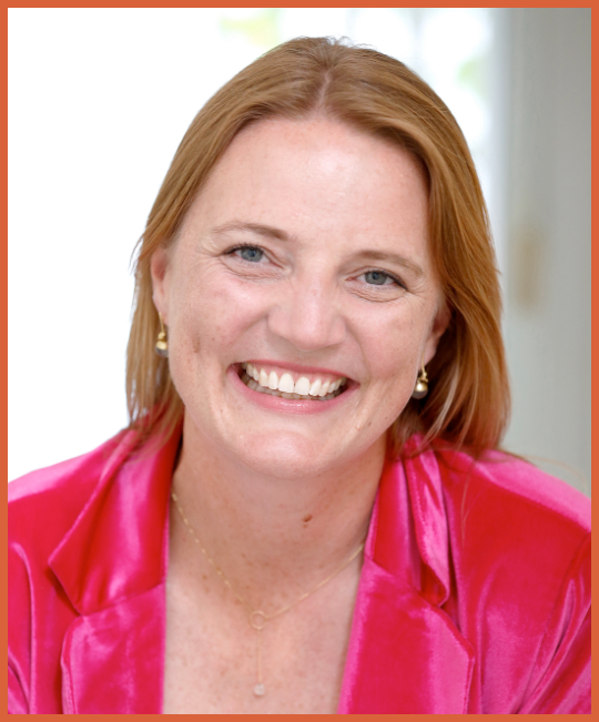 A woman with red hair, blue eyes, and fair skin smiling, wearing a pink jacket and pearl earrings, in a bright indoor setting.