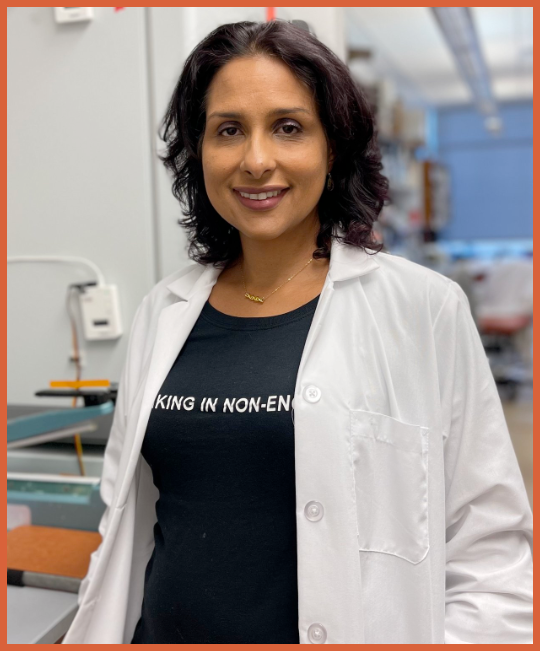 A woman wearing a white lab coat and black t-shirt with the partially visible text "KING IN NON-EN" is smiling and standing in a laboratory or medical setting.