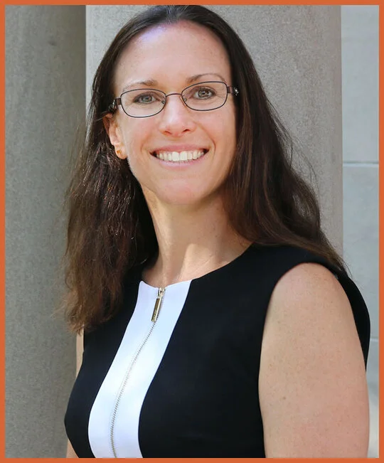 A woman with long brown hair, glasses, and a smile, wearing a sleeveless black and white top, standing outdoors near a building.