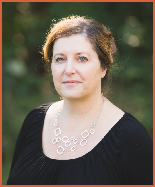 A woman with light skin and brown hair standing outdoors with a blurred green background, wearing a black top and a silver geometric necklace.