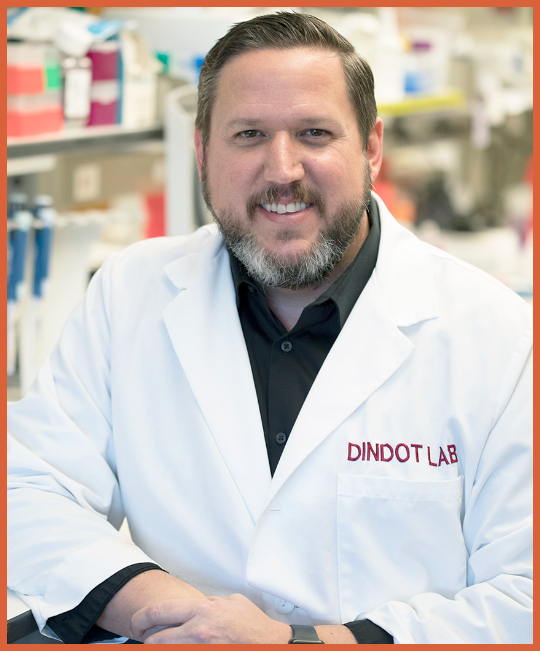 A smiling man with a beard in a laboratory, wearing a white lab coat with "DINDOT LAB" embroidered on it.