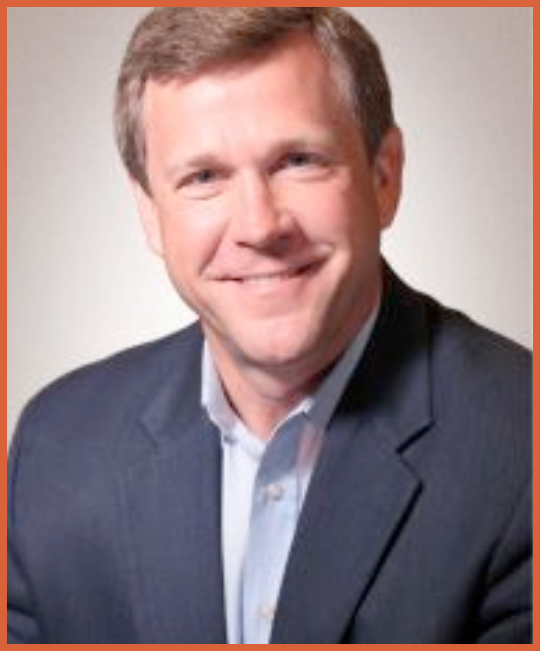 Headshot of a smiling man in a dark suit and light shirt, with short light brown hair, against a neutral background.