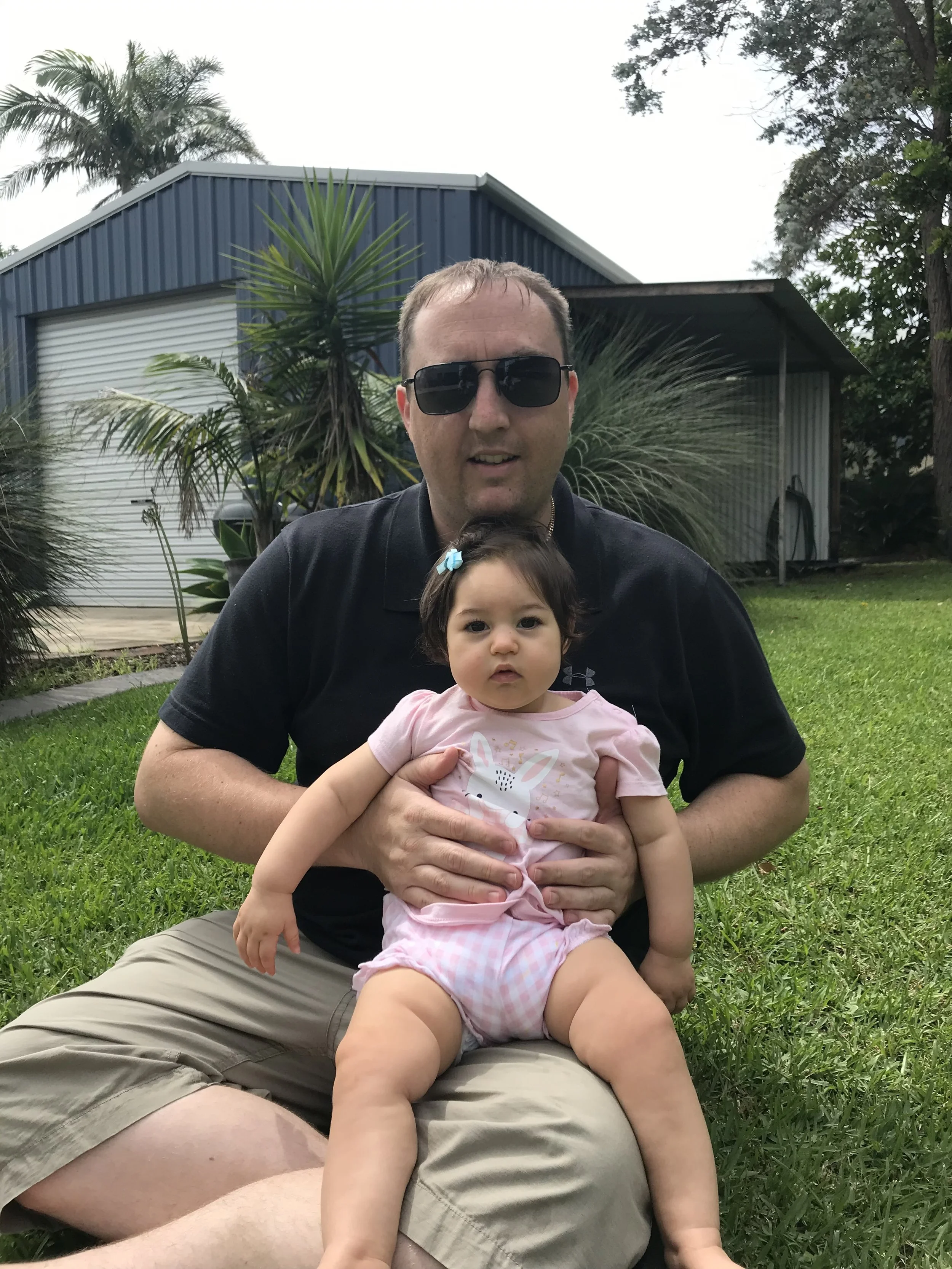 A man with sunglasses and a young girl with FOXG1 syndrome sitting on his lap outdoors on a grassy lawn, with a blue building and trees in the background.