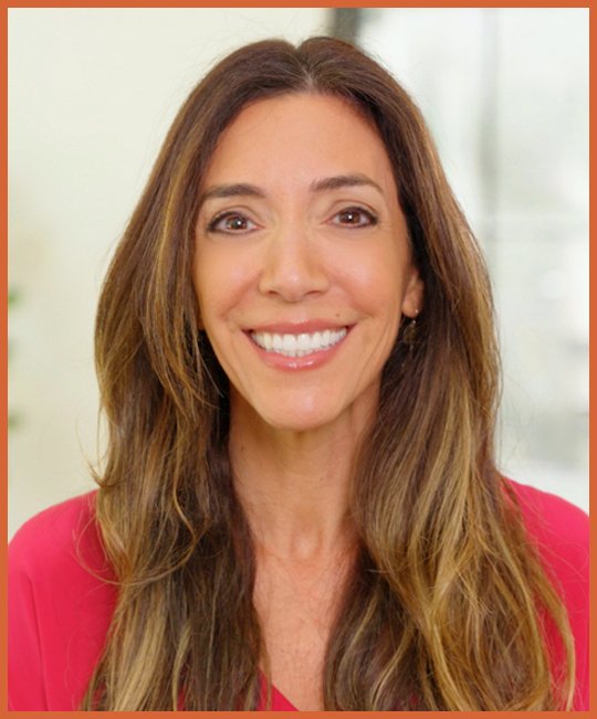Image of Nicole Zeitzer Johnson - cofounder of the FOXG1 Research Foundation  with long, wavy brown hair and a bright smile, wearing a red top, standing in a well-lit indoor space.