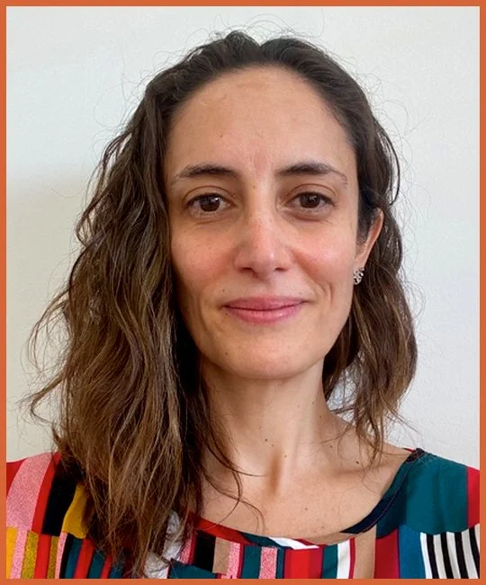 Image of Magdelena Labbe, Secretary of the FOXG1 Research Foundation with wavy brown hair wearing a multicolored top with vertical stripes, standing against a plain light background.