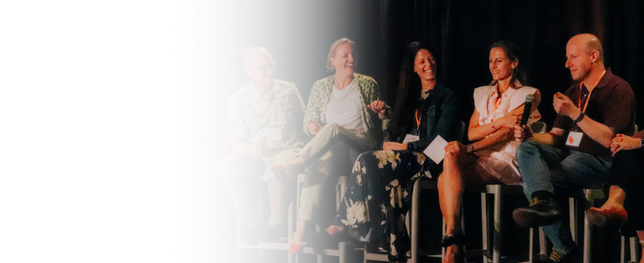 Group of five people sitting on a stage, engaged in a panel discussion, with one person holding a microphone and all smiling.