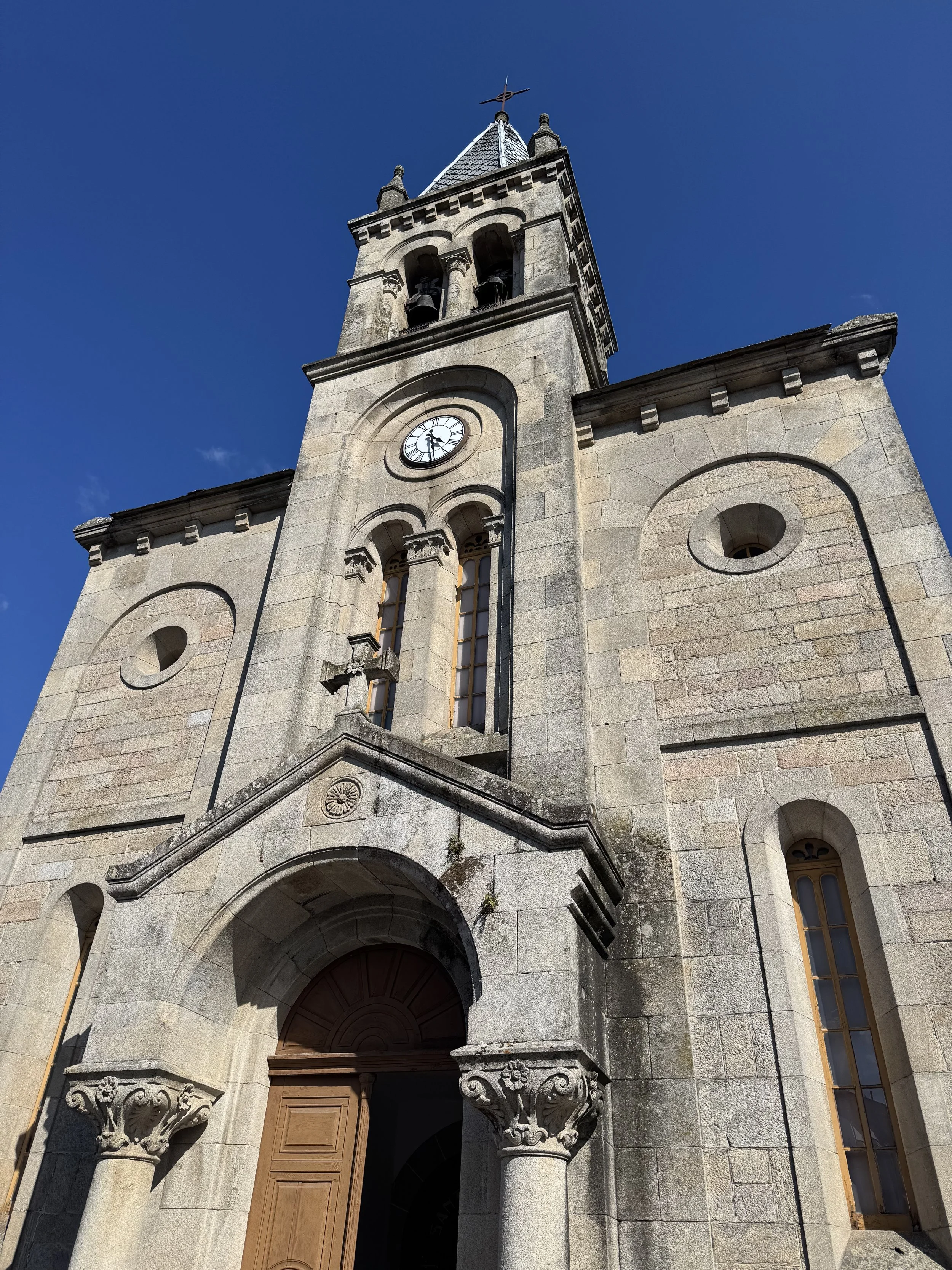 Low-angle view of a historic stone church with a clock tower and a pointed roof, set against a clear blue sky.