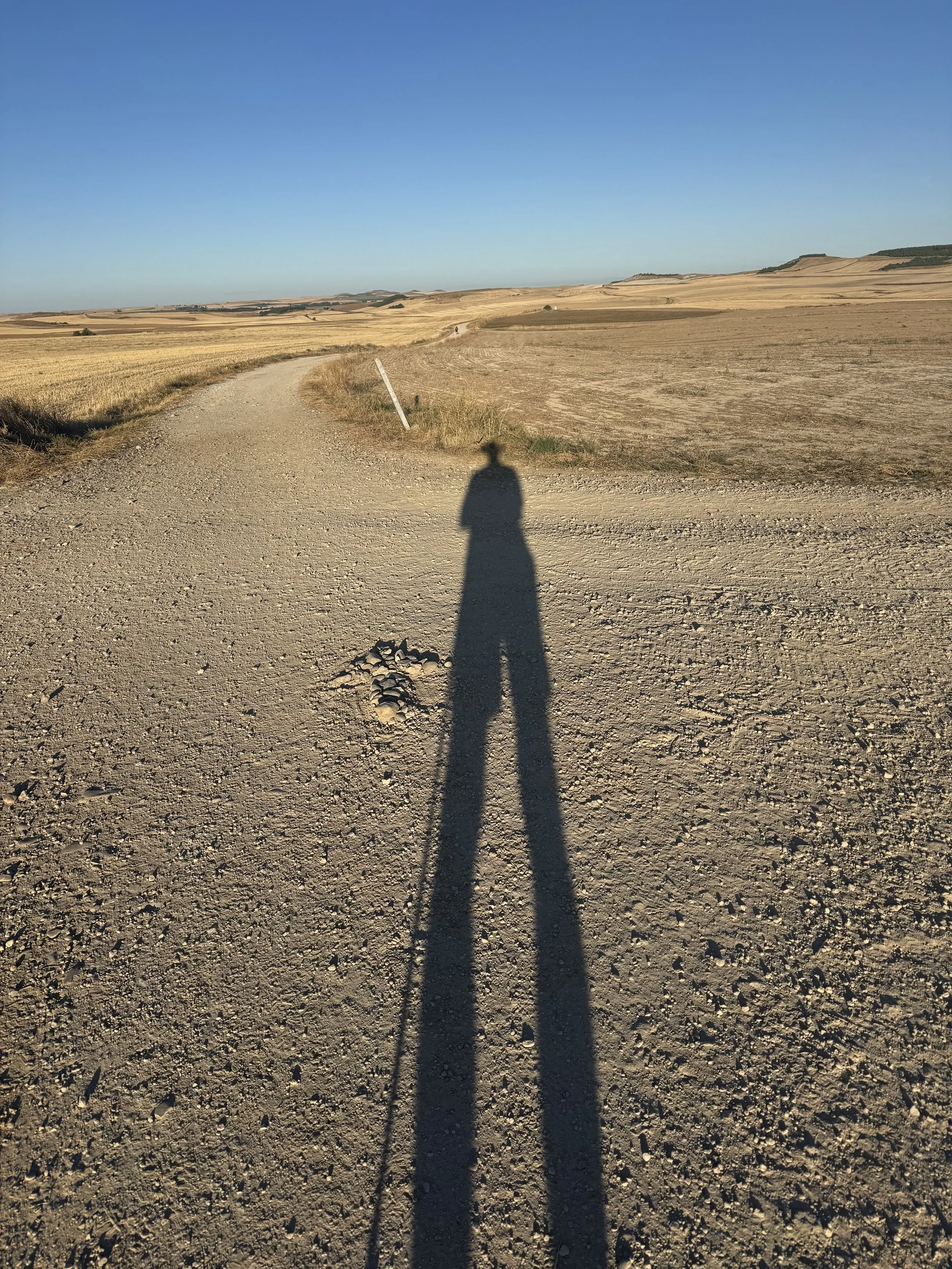 Long shadow of a person wearing a hat on a dirt road in a dry, open landscape under a clear blue sky.