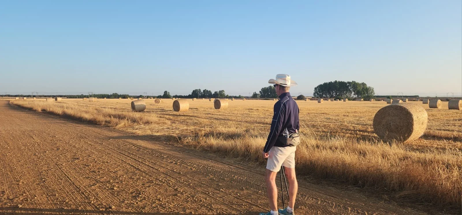 A man in a cowboy hat and shorts standing on a dirt road in a wheat field with hay bales and trees in the background during sunset.