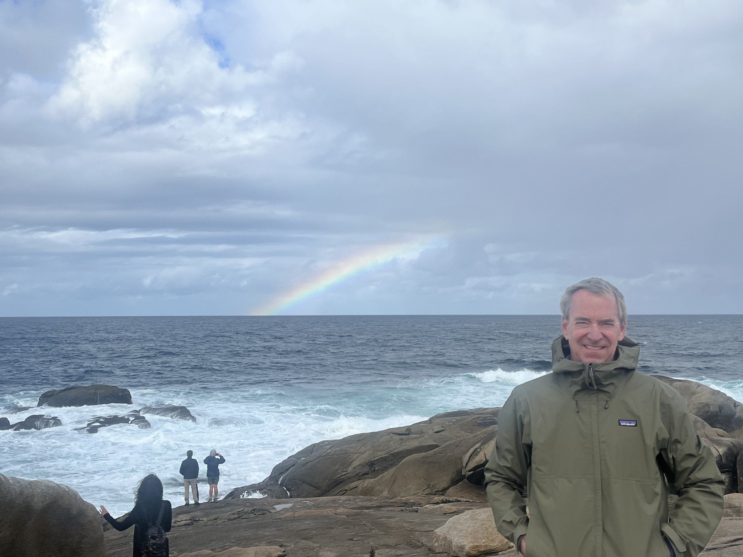 A man in a green jacket standing on rocks near the ocean, smiling at the camera, with three people and a rainbow in the cloudy sky in the background.