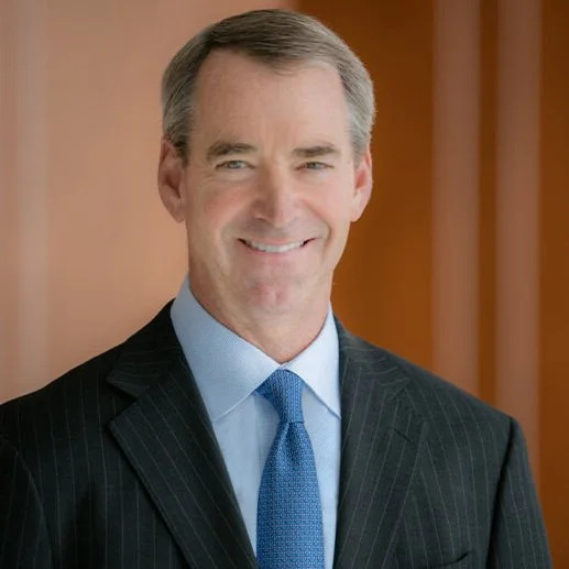 A middle-aged man with gray hair smiling, wearing a dark pinstripe suit, light blue dress shirt, and a blue tie, standing in front of a wooden panel background.