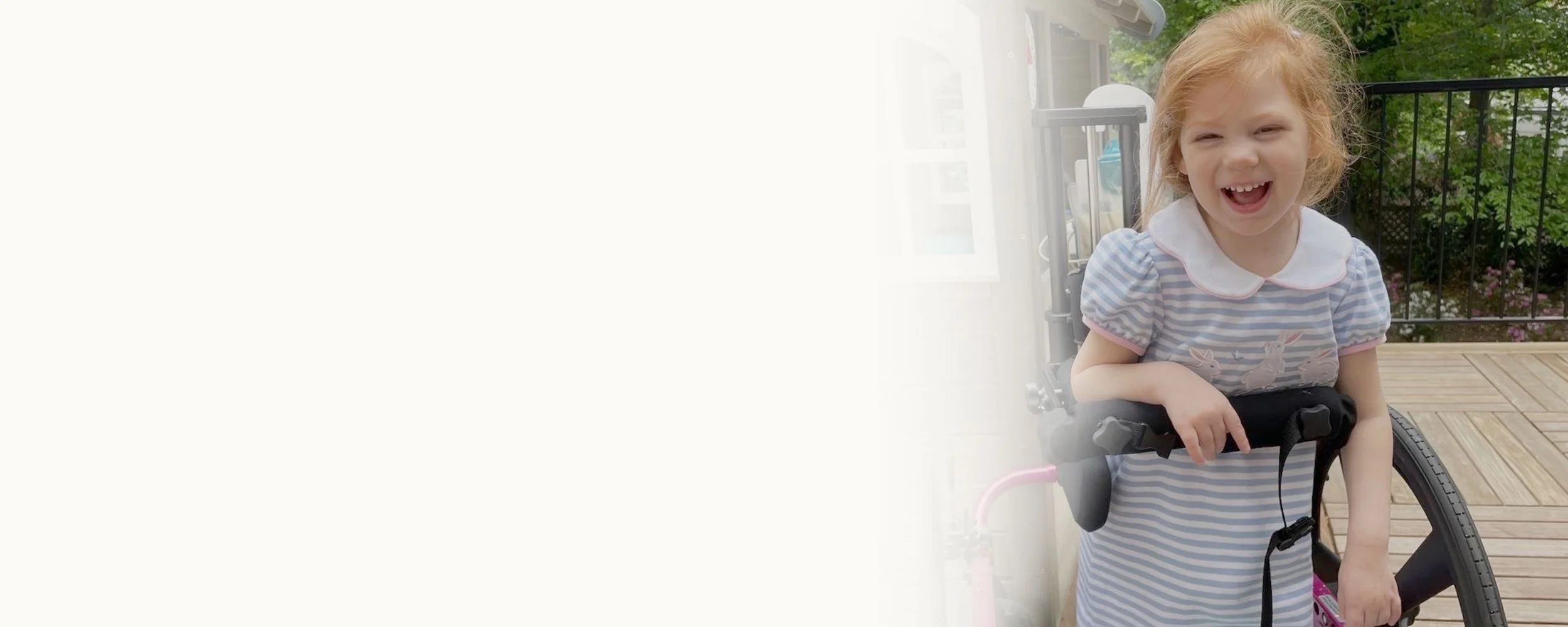 A young girl with red hair smiling and howling outside on a wooden deck, next to a wheelchair, with a black railing and green trees in the background.