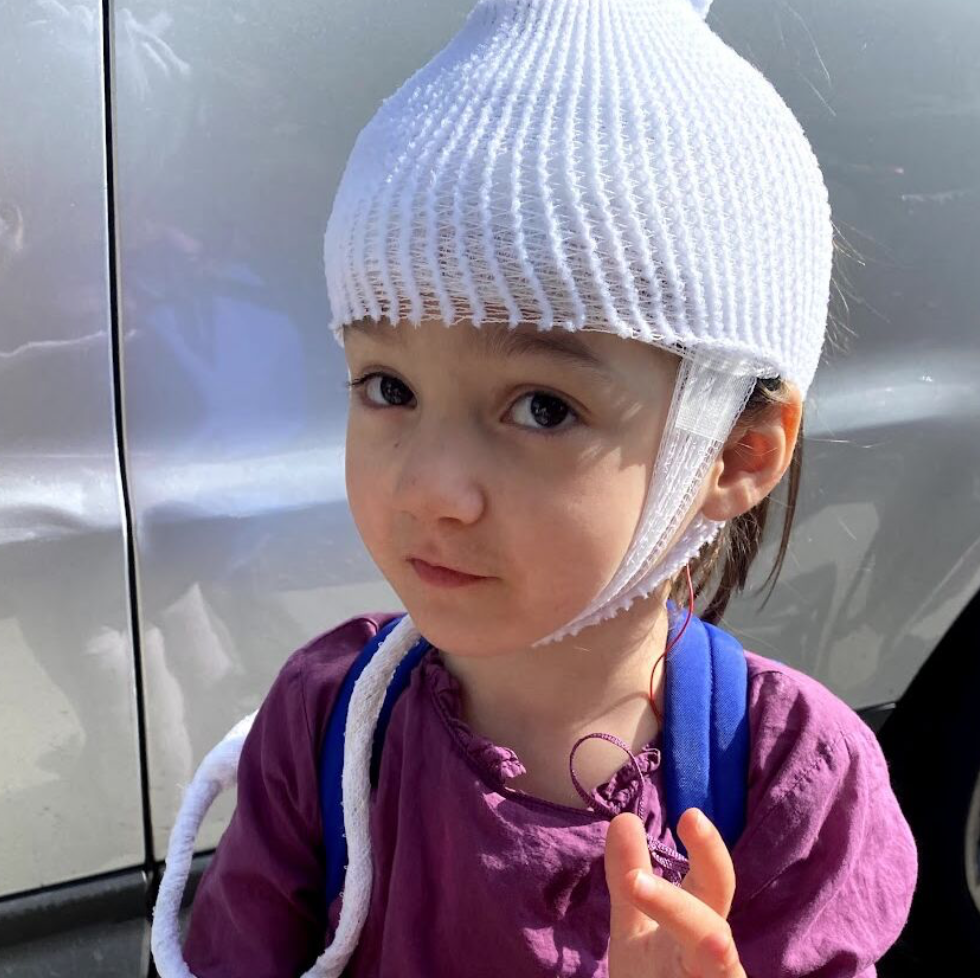 A young girl with light brown hair and light skin wearing a white head wrap and a purple jacket, standing outdoors next to a gray vehicle.