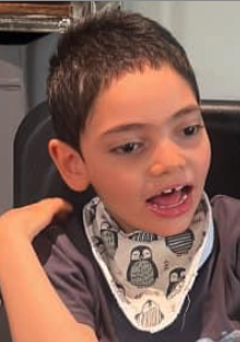 Young boy with short dark hair, wearing a gray shirt and a patterned bib, sitting in a classroom or similar setting.
