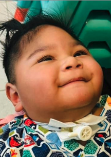 Close-up of a smiling young child with dark hair, wearing a colorful patterned shirt, against a green background.