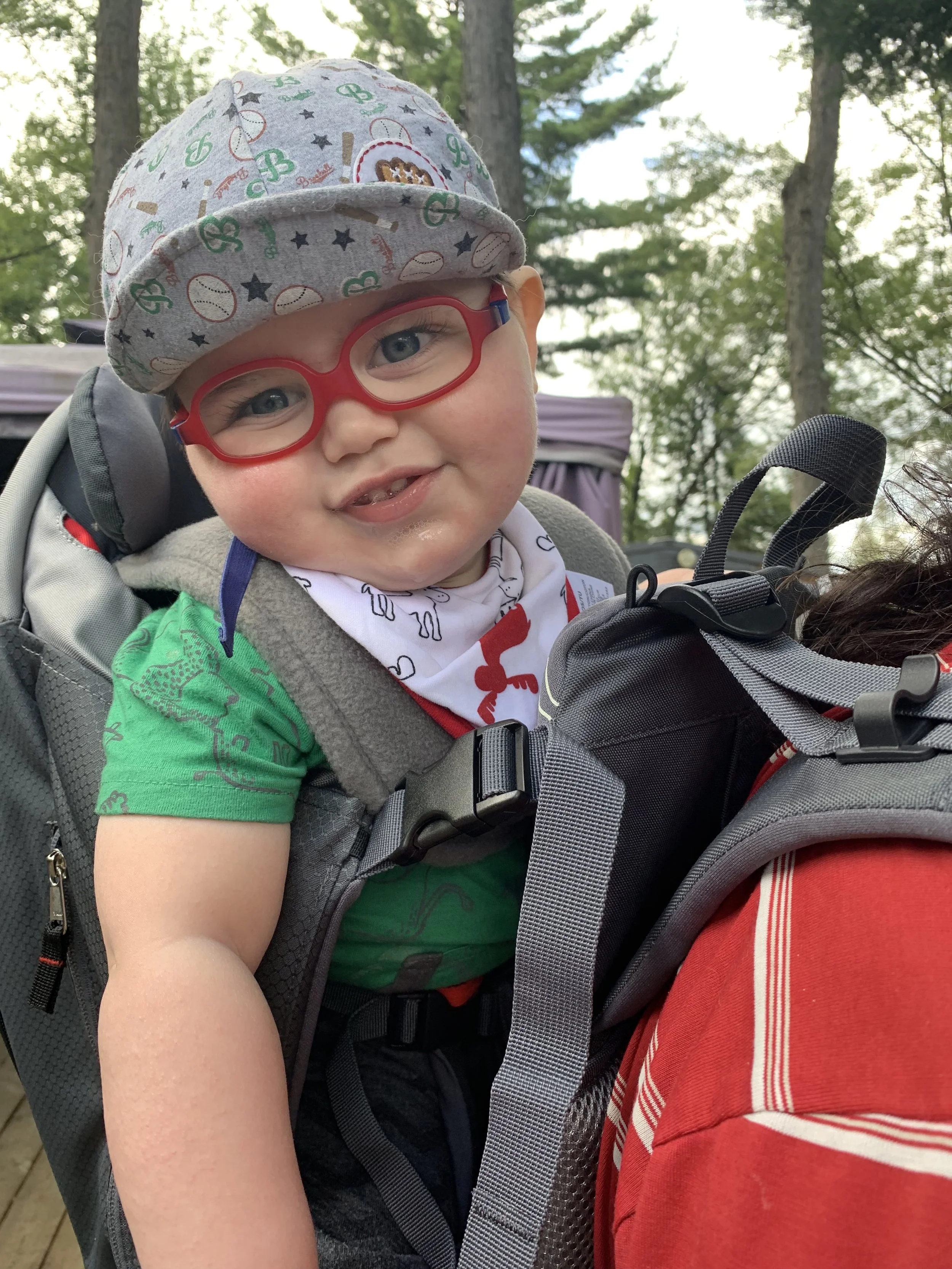 A young child with blue eyes, wearing red glasses, a gray cap decorated with sports and animal patterns, and a white bandana with red and black designs, sitting in a backpack carrier outdoors among trees.