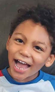 A young boy with curly hair smiling widely and showing his teeth.