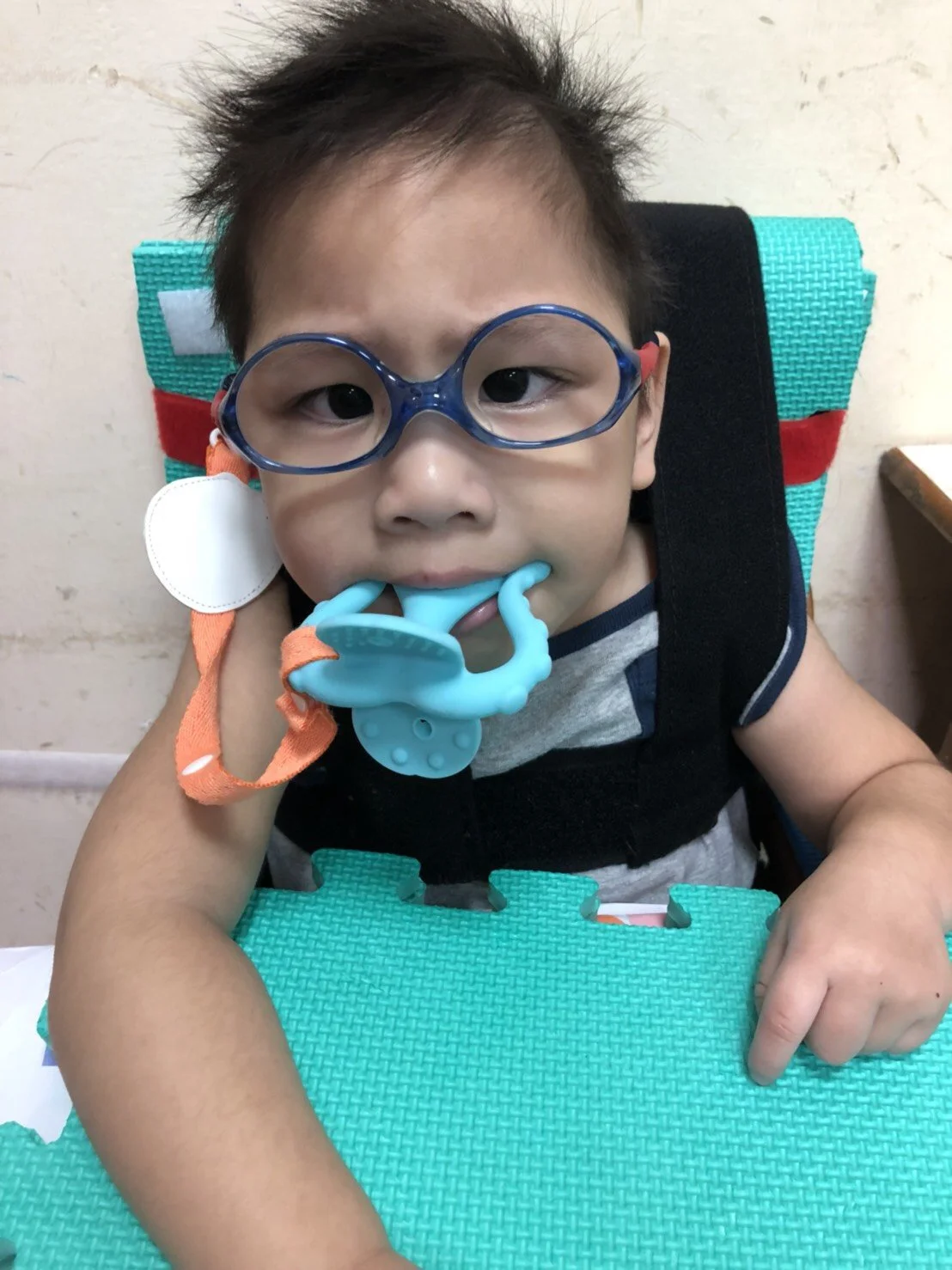 A young boy sitting in a chair, wearing blue glasses, with a pacifier in his mouth, and leaning on a turquoise foam mat.