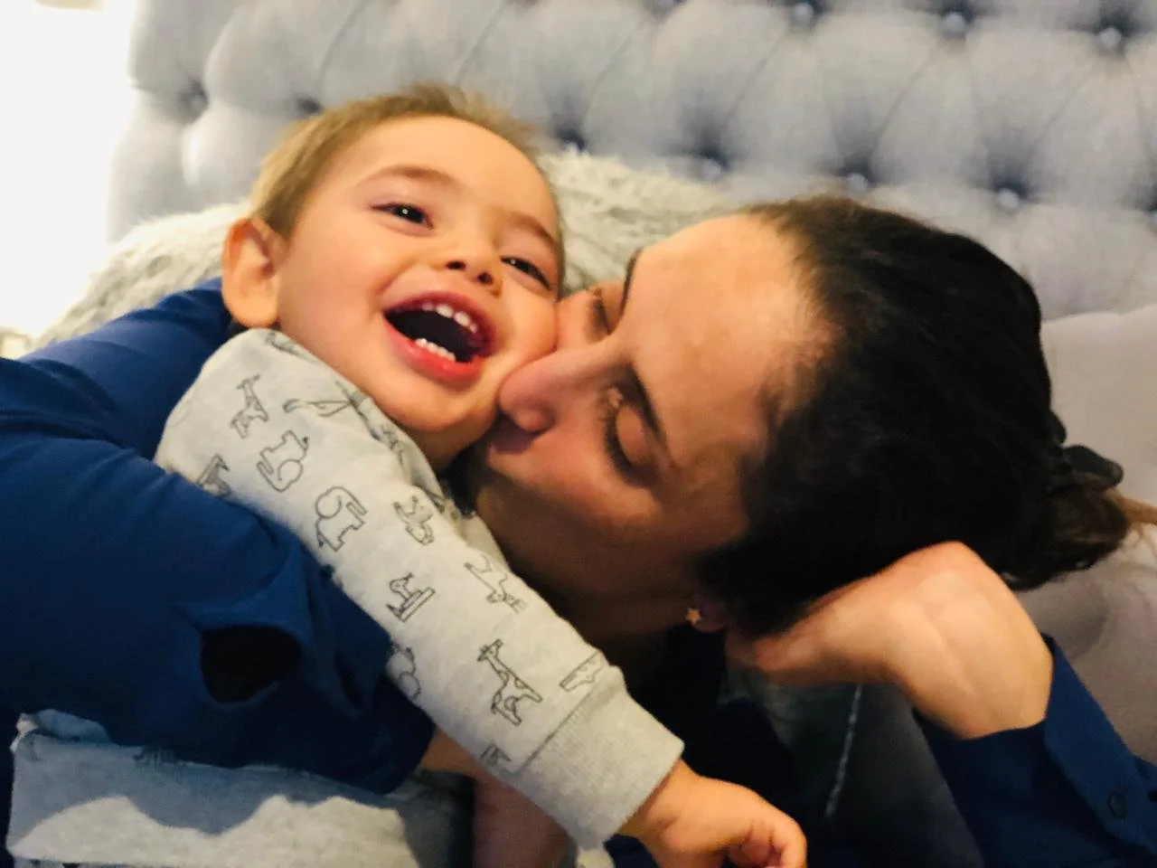 A happy young boy smiling as he hugs a woman, possibly his mother, who is kissing his cheek. They are lying on a bed with a tufted headboard in the background.