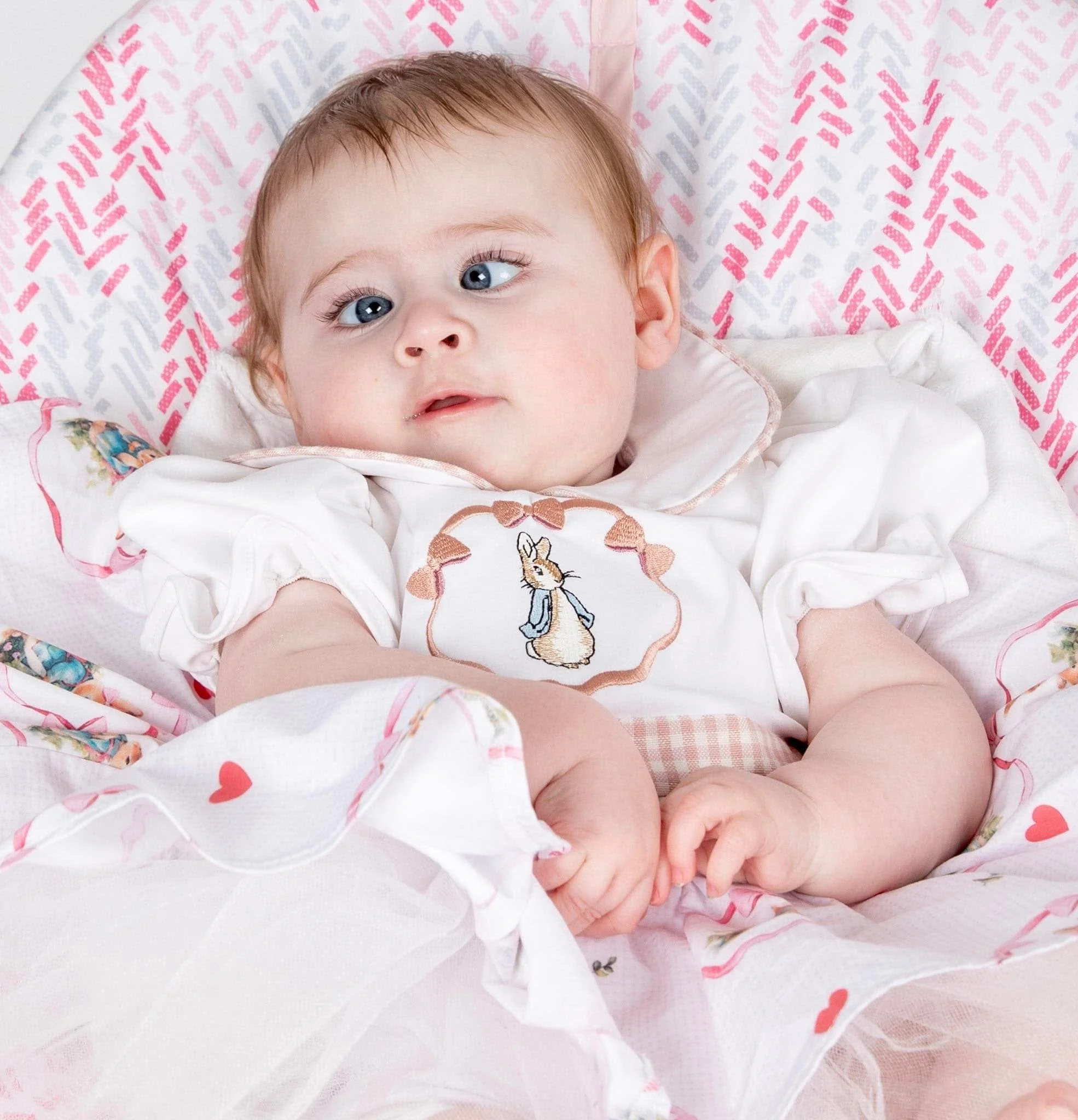 A baby with blue eyes and light brown hair, wearing a white outfit with a bunny embroidery, lying in a decorated crib with pink and white patterned bedding.