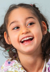 A young girl with a big smile, showing her teeth, with dark hair tied back and wearing a floral shirt.