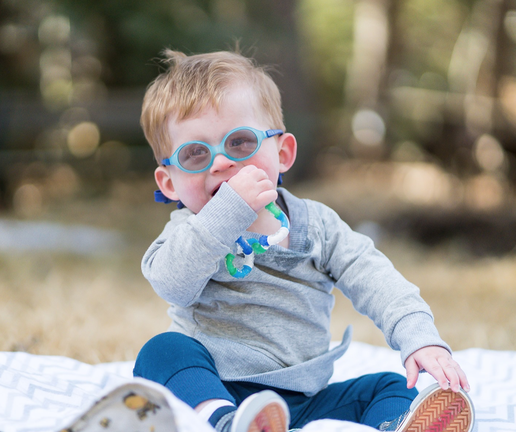 A young boy with blonde hair and blue goggles sitting outdoors on a blanket, wearing a gray hoodie and blue pants, holding a teething toy in one hand and touching his mouth with the other.