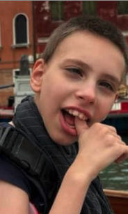 Close-up of a boy with short dark hair, smiling with his mouth open, holding his hand near his chin, outdoors near water and brick buildings.
