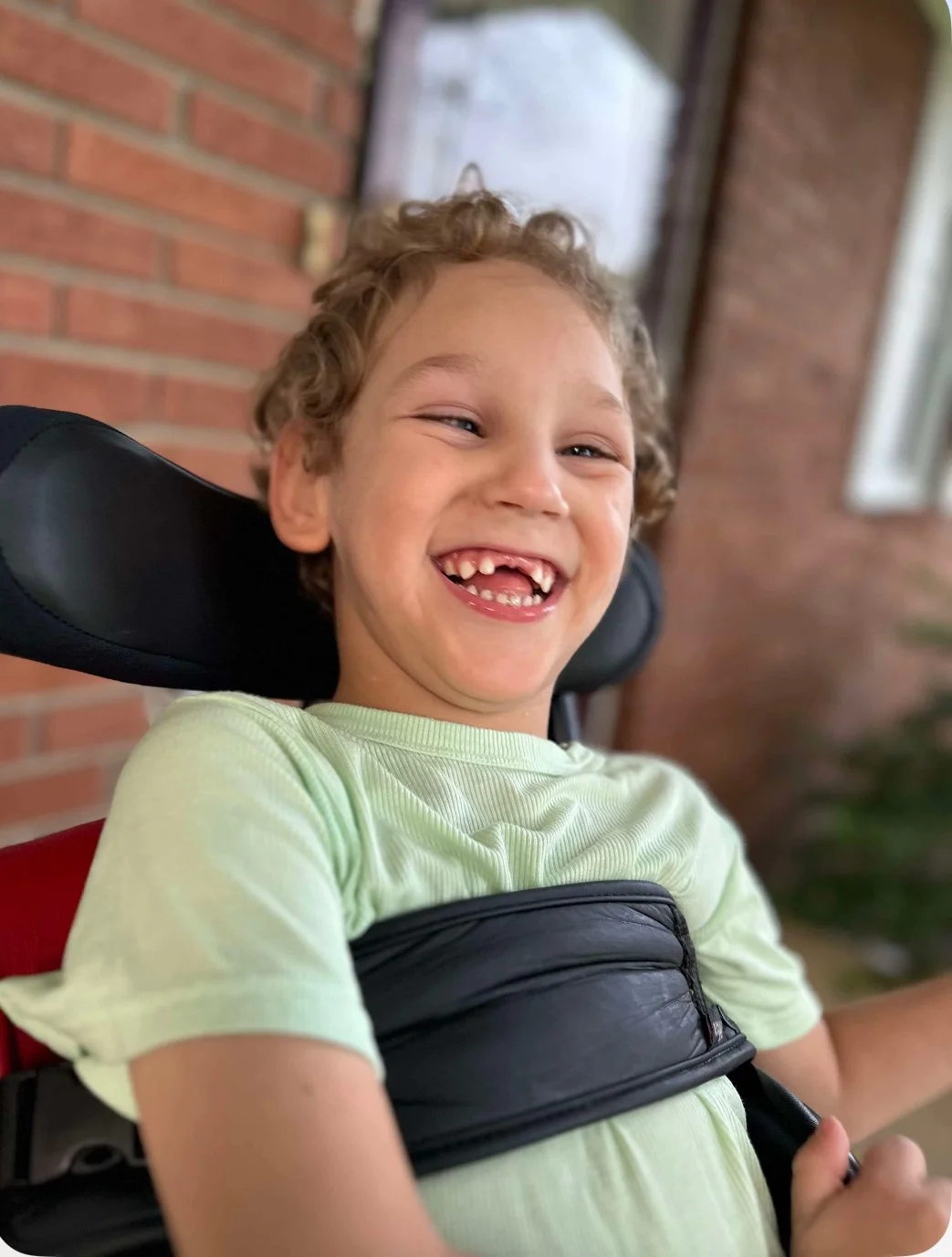 Smiling young boy with curly hair in a green t-shirt sitting on a wheelchair, outdoors in front of a brick wall.
