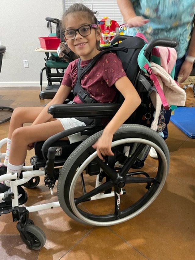A smiling girl in glasses sitting in a motorized wheelchair indoors, with toys and a person standing nearby.