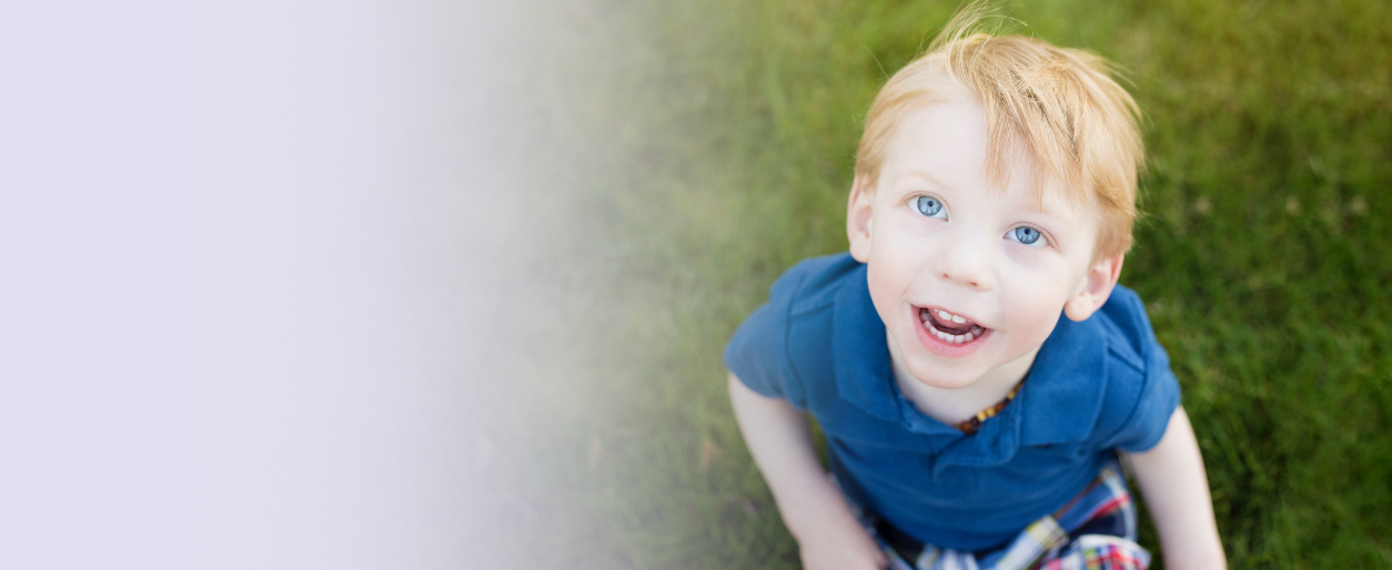A young red-haired boy who has FOXG1 syndrome with blue eyes sitting on green grass, looking up and smiling.
