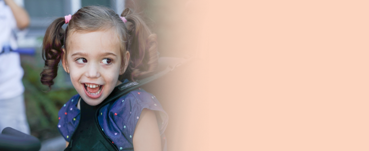 A young girl with pigtails smiling and looking to her side, outdoors, with other children in the background.