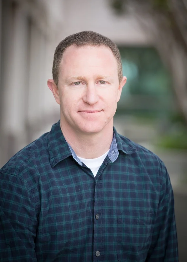 A man with short brown hair and wearing a dark green and blue plaid shirt standing outdoors during daytime.