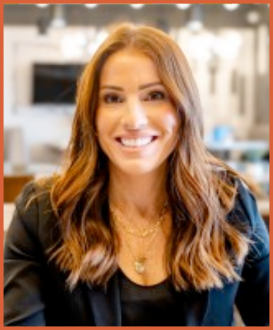 A woman with long, wavy reddish-brown hair smiling at the camera, wearing a black top and blazer, in a bright, modern indoor setting.